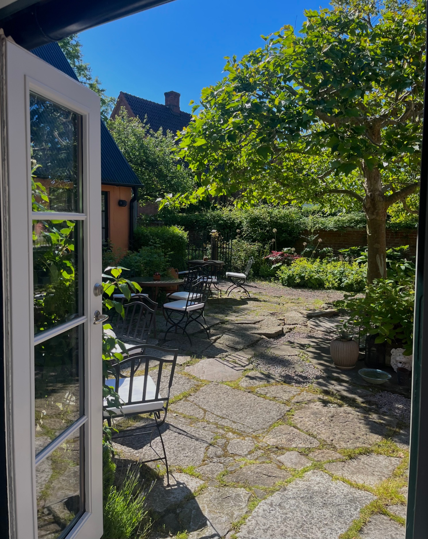 Utsikt från Karnelund genom öppna dörrar till den soliga stengården med trädgårdsmöbler, gröna träd och blå himmel